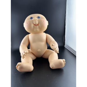 Könnte beinhalten: Eine Vintage-Cabbage-Patch-Kid-Puppe mit einem weichen, fleischfarbenen Vinylkörper. Die Puppe hat ein rundes Gesicht mit rosigen Wangen, blauen Augen und einem lächelnden Ausdruck. Sie sitzt mit ausgestreckten Beinen und leicht ausgestreckten Armen.