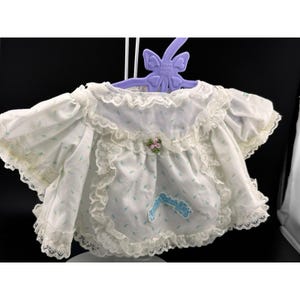 May include: A white doll dress with blue floral patterns and lace trim. The dress has short sleeves and a ruffled collar. A small pink flower and blue ribbon accent the front. The dress is hanging on a purple hanger.