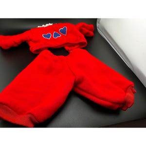 May include: A bright red two-piece outfit consisting of a long-sleeved top and matching pants. The top features a white lace collar and three blue heart-shaped appliqués with a white polka dot pattern across the chest area.
