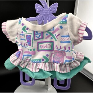 Könnte beinhalten: Ein Vintage-Puppenkleid mit einem weißen Oberteil mit geometrischen Formen in Türkis, Lila und Blau. Das Kleid hat kurze Ärmel mit rosa und weißen Streifen, einen türkisfarbenen Rock und einen Rüschensaum. Das Kleid hängt an einem lila Kleiderbügel mit der Aufschrift "Dress Along Dolly".