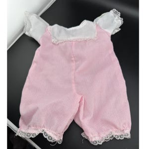 May include: A one-piece romper featuring a pink and white gingham pattern on the lower section and a solid white bodice. The garment includes short puff sleeves, a square neckline, and white lace trim detailing along the collar and leg openings.
