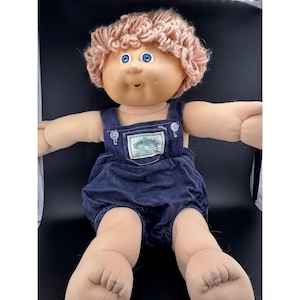 May include: A vintage Cabbage Patch Kid doll with a peach-colored yarn hairstyle and blue eyes. The doll is wearing a denim overall outfit with a green and white patch on the chest. The doll is sitting with its arms outstretched.