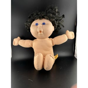 May include: A vintage baby doll with a soft, flesh-toned body and a round face. The doll has blue eyes, a small open mouth, and a head of dark, curly hair. Its arms are outstretched, and it is sitting with its legs bent.