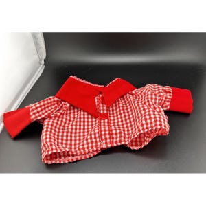 May include: A red and white gingham shirt with a red collar and cuffs. The shirt features a button closure and is likely designed for a doll or small toy. The material appears to be cotton or a similar fabric.
