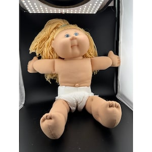 May include: A vintage Cabbage Patch Kid doll with blonde yarn hair and blue eyes. The doll is wearing a white diaper and has its arms outstretched. The doll is tan-skinned and has a soft, plush appearance.