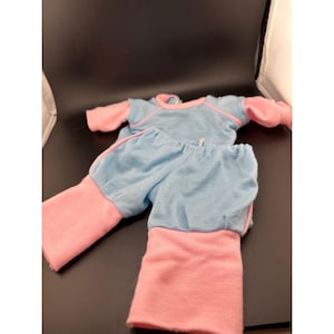 May include: A two-piece doll outfit in light blue and pink. The long-sleeved top and pants have pink cuffs and trim. The outfit is made of a soft, possibly knit, material.