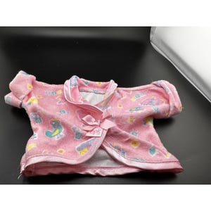 May include: Pink doll-sized pajama top with a satin trim and a bow closure. The top is decorated with colorful cartoon images of birds, clouds, and other shapes. The tag reads "cpk".