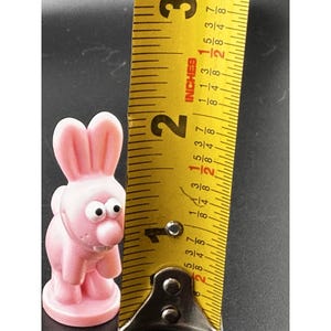 May include: A pink bunny figurine with large eyes and a prominent nose stands next to a yellow measuring tape. The bunny is approximately 2 inches tall. The tape measure shows inches.