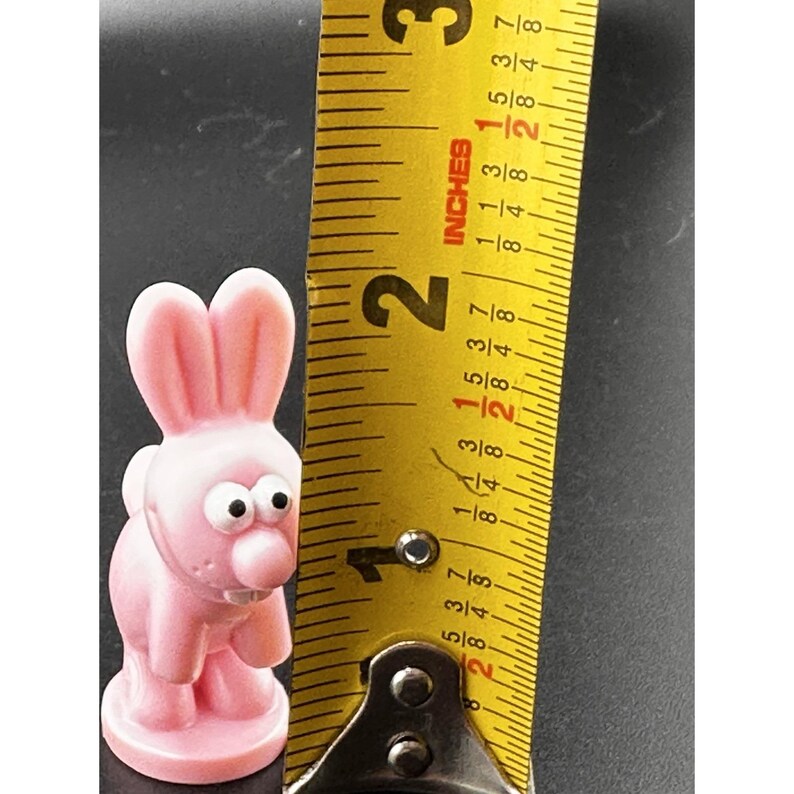 May include: A pink bunny figurine with large eyes and a prominent nose stands next to a yellow measuring tape. The bunny is approximately 2 inches tall. The tape measure shows inches.