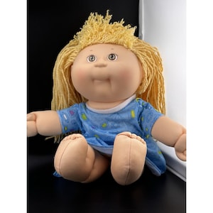 Könnte beinhalten: Eine Vintage Cabbage Patch Kid Puppe mit blondem Garnhaar und einem blauen gemusterten Kleid. Die Puppe hat einen weichen, plüschigen Körper und sitzt mit ausgestreckten Beinen. Die Augen der Puppe sind hellbraun.