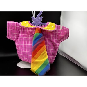 May include: A pink plaid shirt with a yellow polka dot collar and a rainbow tie. The shirt is hanging on a purple hanger with a bow design. The shirt is designed for a doll.