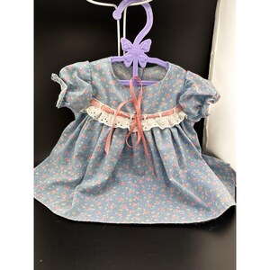 May include: A blue floral print dress with short sleeves and white lace trim. The dress is suspended on a purple butterfly-shaped hanger and has a pink ribbon tied around the waist.