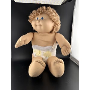Puede incluir: Una muñeca Cabbage Patch Kid vintage con piel bronceada, ojos azules y cabello rubio rizado. La muñeca lleva un pañal con detalles amarillos y está sentada con las piernas dobladas. La muñeca tiene una expresión sonriente.