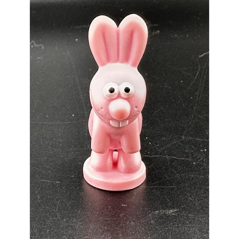 May include: A pink bunny figurine with large ears, white eyes, and a round pink nose. The bunny has a small mouth with two visible teeth and stands on a circular base. The figurine is made of a smooth, glossy material.