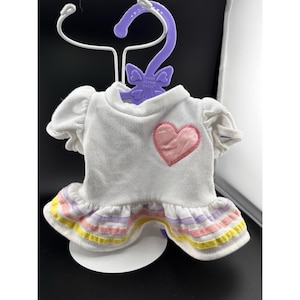 May include: White doll dress with a pink heart applique and ruffled hem. The hem features horizontal stripes in pastel colors of yellow, pink, and lavender. The dress is displayed on a purple hanger with the text "Dress Along Dolly".