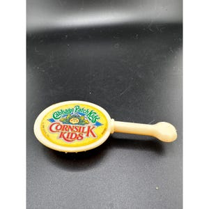 May include: A vintage Cabbage Patch Kids Cornsilk Kids toy. The toy is a light yellow color with an oval-shaped head and a handle. The head features the Cabbage Patch Kids logo and the words "Cornsilk Kids" in a colorful design.