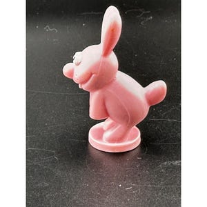 May include: A pink plastic bunny figurine with a smiling face, long ears, and a small tail. The bunny is standing on a circular base. The figurine is a solid, smooth color.