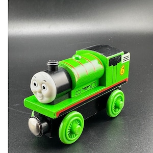 May include: A green wooden toy train with a smiling face, black accents, and the number 6 on the side. The train has green wheels and a black base. The train is a classic toy.