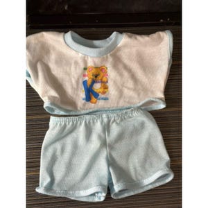 May include: A light blue and white doll outfit. The top is white with a light blue trim and a cartoon bear graphic with the letter "K". The matching shorts are light blue.