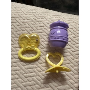 May include: A yellow heart-shaped rattle with a bow detail, a purple barrel-shaped container with the text CPK embossed on the side, and a yellow heart-shaped pacifier with the text CPK embossed on the handle, all made of plastic.