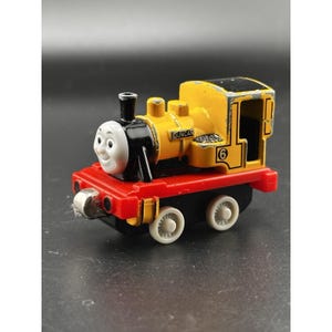 Op de afbeelding: Een gele en zwarte speelgoedtrein met de naam "Duncan" aan de zijkant. De trein heeft een rode basis, zwarte wielen en het nummer "6" is zichtbaar. De trein heeft een lachend gezicht.