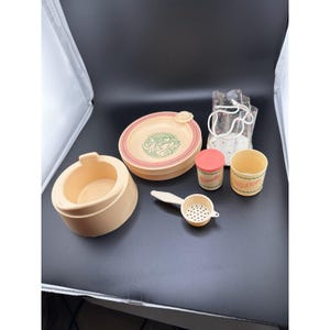 Può includere: Una collezione di articoli per l'infanzia vintage color pesca chiaro. Include una ciotola rotonda con un design rosa e verde, un piccolo contenitore con l'etichetta "Polvere", una tazza con l'etichetta "Cotone", un colino e un piccolo contenitore con coperchio.