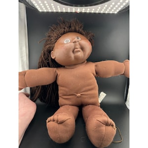 May include: A dark-skinned doll with a soft fabric body and brown yarn hair. The doll features a round, molded plastic face with large, light-colored eyes, a small nose, and a closed mouth. The arms and legs are extended outward from the torso.