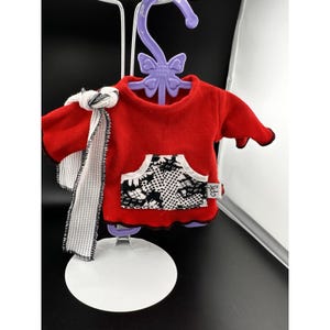 May include: A red doll-sized hoodie with a black and white patterned pocket and black trim. A white and black patterned scarf is draped over the shoulder. The hoodie is hanging on a purple hanger that says "Dress Along Dolly".