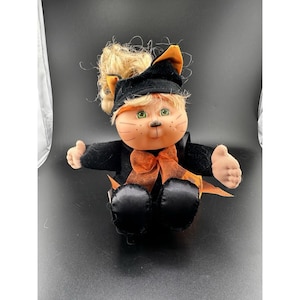Peut inclure: Poupée en peluche déguisée en chat noir avec un nœud et un chapeau orange. La poupée a des cheveux blonds, des yeux verts et un visage peint avec des moustaches. La poupée est assise, les bras tendus.