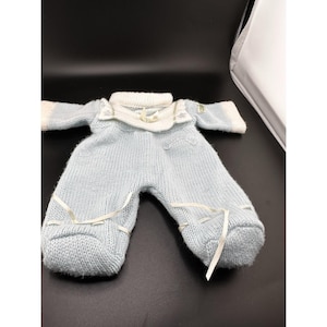 Könnte beinhalten: Hellblaues Baby-Strick-Outfit mit weißer Verzierung und Bändern. Das Outfit besteht aus einem langärmeligen Oberteil und einer Hose mit Füßen. Der Kragen und die Manschetten sind weiß, und das Outfit ist mit weißen Bändern gebunden.