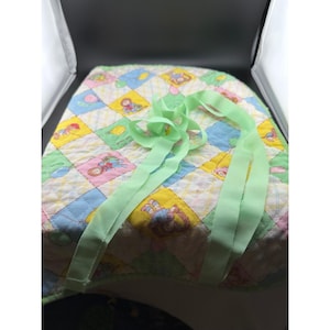 May include: A quilted baby changing pad with a colorful patchwork design featuring cartoon characters. The pad has a light green ribbon attached. The patchwork includes yellow, blue, pink, and green squares.