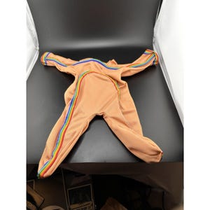 Könnte beinhalten: Pfirsichfarbener Overall mit Regenbogenstreifen an Ärmeln und Beinen. Der Overall hat einen Reißverschluss vorne und besteht aus einem weichen Rippstoff. Die Regenbogenstreifen umfassen Rot, Orange, Gelb, Grün, Blau und Lila.
