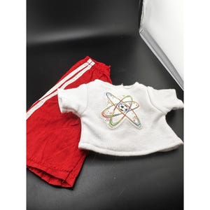 May include: A white short-sleeved t-shirt featuring a graphic of a soccer ball surrounded by colorful orbital rings and small stars, paired with red athletic shorts that have two vertical white stripes running down the side of the leg.