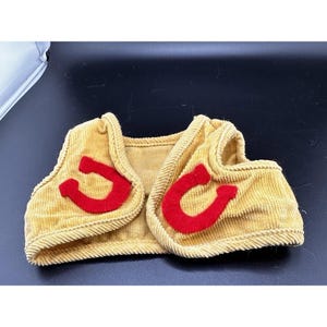 Op de afbeelding: Een mosterdgele corduroy vest met rode hoefijzer applicaties. Het vest heeft een ronde halslijn en een eenvoudig, open ontwerp. De hoefijzer applicaties zijn felrood van kleur en zijn aan de voorkant van het vest geplaatst.