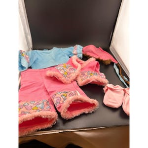 May include: A collection of doll clothes in various shades of pink and blue. The set includes a pink jacket, pants, and mittens, all trimmed with fluffy pink material. A blue shirt and a pink hat with a brown fur trim are also included.