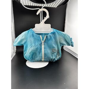 Puede incluir: Una chaqueta de color azul claro con mangas cortas, cremallera frontal blanca y puños y dobladillo elásticos. La prenda presenta el texto Cabbage Patch Kids en letras de color verde oscuro en el lado derecho y se muestra en una percha blanca.