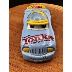 May include: A vintage blue Tonka Motorsports toy car with yellow accents. The car features the words "Tonka" and "Team Tonka Motorsports" in red. The toy car is on a wooden surface.
