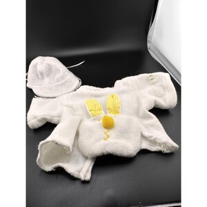 May include: A white terry cloth baby outfit with a matching hat. The outfit is designed to look like a bunny, with yellow ears and a yellow pom-pom nose. A yellow zig-zag stitch forms the mouth.