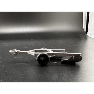 Könnte beinhalten: Ein silberner Spielzeuganhänger mit schwarzen Rädern. Der Anhänger hat eine Deichsel vorne und eine Ladefläche mit einem Metallrahmen.