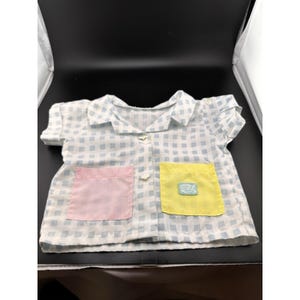 May include: A short-sleeved, collared shirt with a blue and white checkered pattern. It features a pink pocket on the left and a yellow pocket on the right. The yellow pocket has a small embroidered design. The shirt has two buttons.