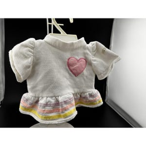 Op de afbeelding: Wit poppen shirt met een roze hart applicatie op de borst. De top met korte mouwen heeft een gefranjerde zoom met horizontale strepen in pastelkleuren roze, geel en lavendel. Het shirt hangt aan een witte hanger.