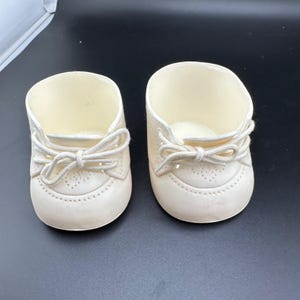 May include: A pair of off-white doll shoes featuring a rounded toe, a lace-up front with matching fabric laces tied in a bow, and a subtle perforated decorative pattern along the toe cap and side panels of the footwear construction.