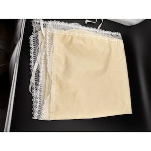 Op de afbeelding: Een gevouwen, lichtgele stof met een delicate witte kanten rand. Het kant is ingewikkeld ontworpen en voegt een vleugje elegantie toe. Witte linten zijn aan de bovenrand bevestigd. De stof lijkt zacht en glad.