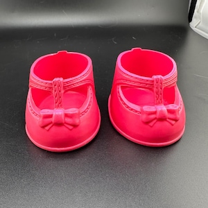 Puede incluir: Un par de zapatos de muñeca de color rosa intenso con punta redondeada, diseño de correa en T y un pequeño lazo moldeado en la parte delantera. Los zapatos tienen una construcción de plástico liso y sólido con costuras en relieve.