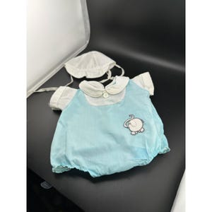 Peut inclure: Une barboteuse bleu clair avec des manches courtes blanches, un col rond blanc et un bouton blanc unique à l'encolure. Un écusson brodé en forme d'éléphant est placé sur le devant. Un bonnet blanc assorti avec des liens est inclus.