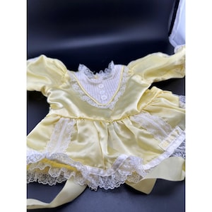May include: A pale yellow satin dress featuring long sleeves, a white pinstriped bodice insert, and three small white buttons. The garment is accented with white lace trim along the neckline, waist, and hem, and includes long satin ties at the waist.
