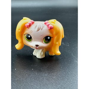 May include: A small plastic dog figurine with a cream-colored face, large green eyes, and long yellow ears styled with pink bows. The toy features a brown nose, a small mouth, and textured white paws, capturing a stylized animal character design.
