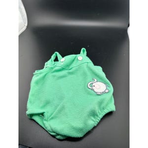 May include: A bright green sleeveless romper featuring two white buttons at the neckline and an embroidered elephant patch on the lower right side. The garment has a rounded silhouette with gathered leg openings and a soft, textured fabric finish.