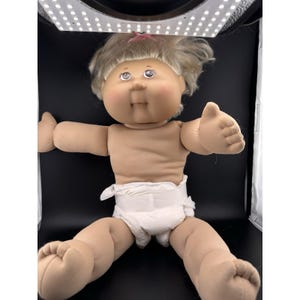 May include: A vintage Cabbage Patch Kids doll with blonde hair and blue eyes. The doll is wearing a white diaper and has its arms outstretched. The doll is made of soft material and has a classic, nostalgic appearance.
