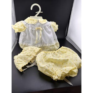 May include: A vintage baby doll outfit set, comprising a white and yellow floral dress, matching bloomers, and a bonnet. The dress features a Peter Pan collar and lace trim. The set is displayed on a white hanger.